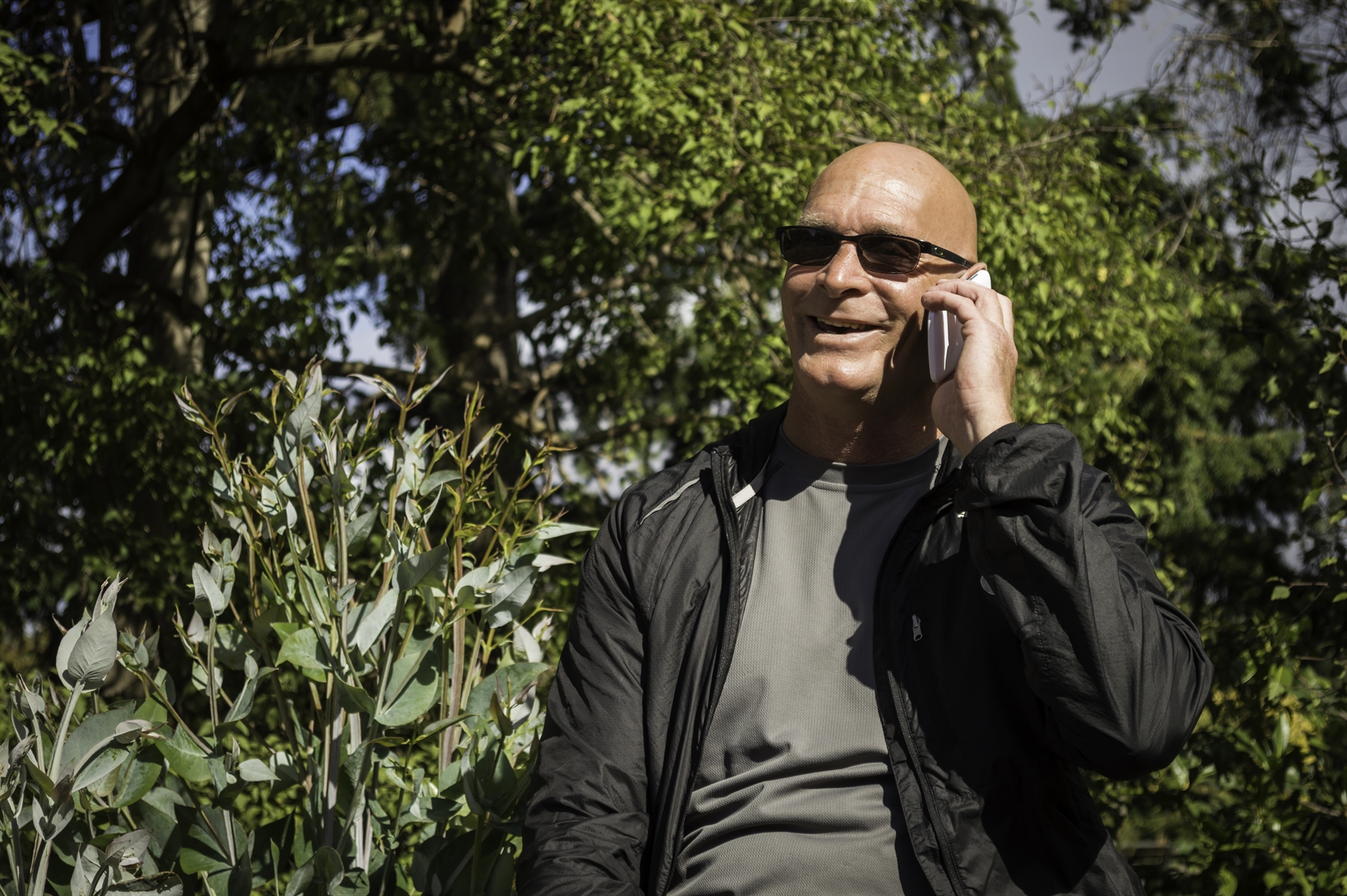A middle-age man is talking on his mobile phone. He is outside amongst trees and is wearing dark glass, a black jacket and a grey t-shirt.