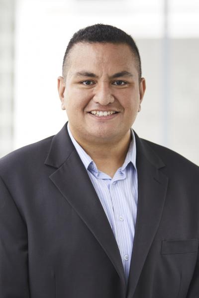Fale Andrew Lesā is smiling and wearing a light blue shirt with a dark blazer.