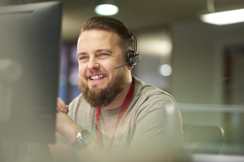 A young man with a beard sitting in front of a computer in an office wearing a telephone headset and red lanyard smiling while talking.