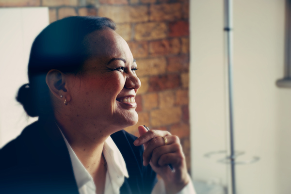 A Māori woman wearing a white shirt and jacket is smiling. Her dark hair is tied in a low tidy bun and she has holding a pen in her left hand.