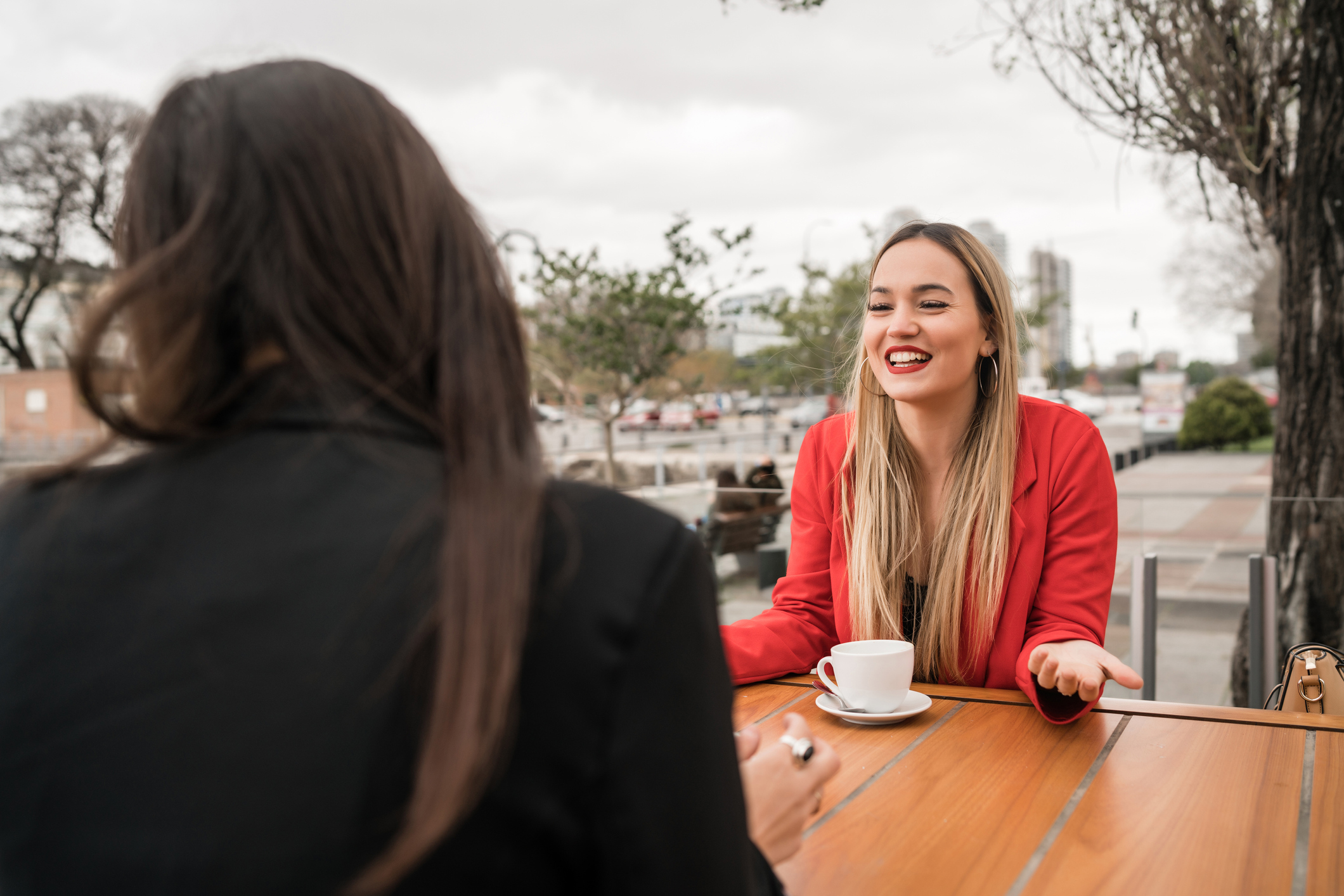 Two young women sitting at a table outside having coffee. The woman facing the camera is smiling at the other woman and wears a tidy red jacket. The second woman sits with her back to the camera and is wearing a black jacket.