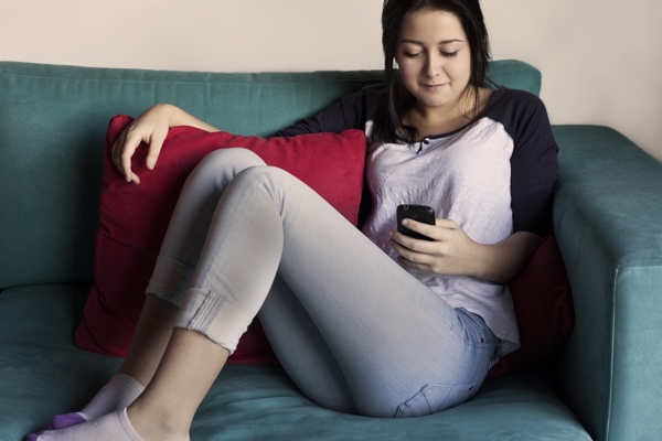 A young woman sitting on a green couch looking down at her phone. She has long dark hair, a white and black sleeved shirt and blue jeans. She sits with her feet up on the couch, and is leaning on a large red cushion.