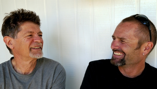 Two men sitting in front of a white wall. From left a man with spiky, dark hair and a short grey beard wearing a light grey t-shirt looking at a smiling man with short hair, and a dark beard with black sunglasses on top of his head wearing a black shirt.